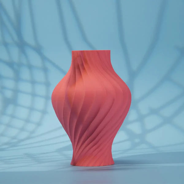 A coral-pink, matte vase with a spiral design—3D printed using Flashforge PLA 1.75mm Matte Colour filament from 3D Printing Perth - Cirrus Link—stands on a blue surface with crisscrossing shadow patterns in the background.