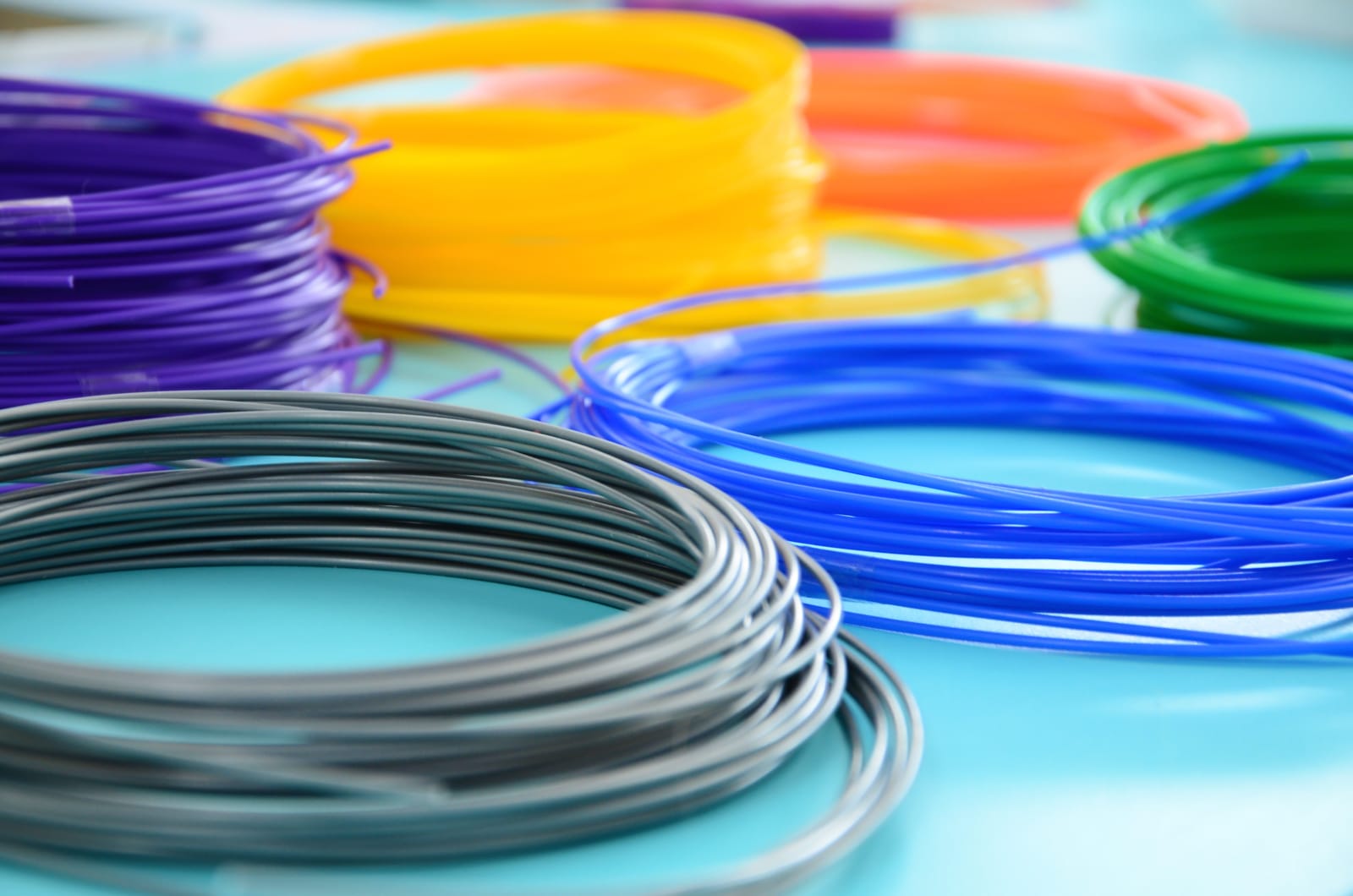 Coils of colored plastic filament, including gray, blue, green, yellow, orange, and purple, arranged on a light blue surface.