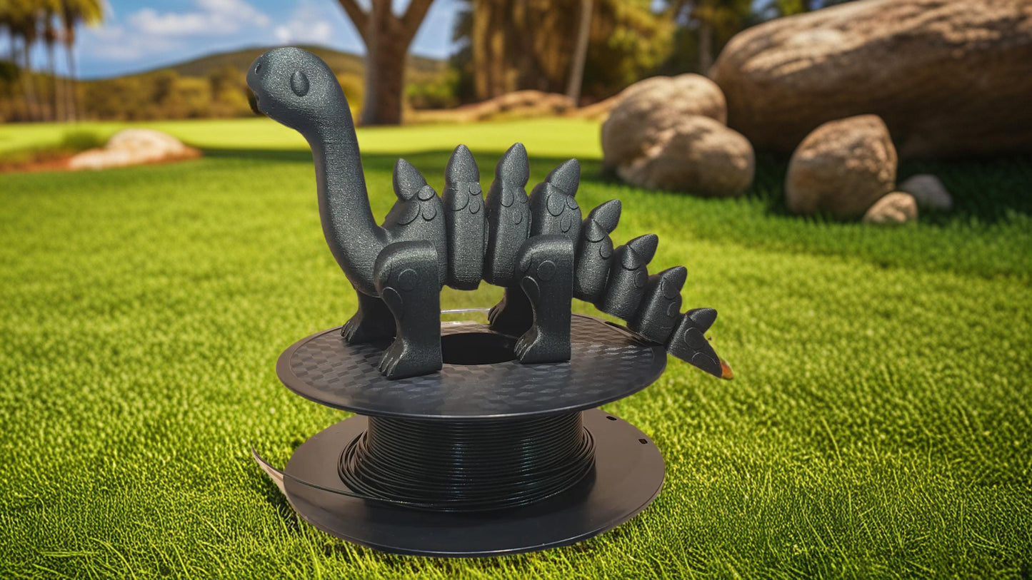 A 3D-printed dinosaur, made from Hello3D PLA Magic Colour 1.75mm filament, sits on a spool of black filament atop green grass, with rocks and trees in the background.
