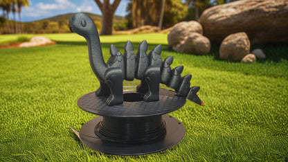 A 3D-printed dinosaur, made from Hello3D PLA Magic Colour 1.75mm filament, sits on a spool of black filament atop green grass, with rocks and trees in the background.