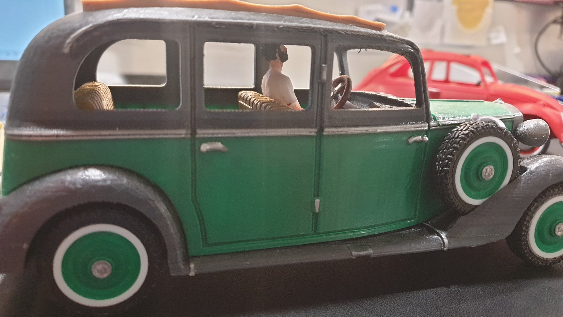Side view of a miniature green STL File : 1930 Mercedes Vehicle by Gordon Nezich with a driver inside. A red classic car model is visible in the blurred background.