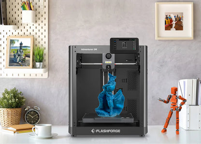 A FlashForge 3D Printer FlashForge Adventurer 5M - High-Speed Direct Extruder on the desk is printing a blue figure with high printing speed. The workspace features a plant, a figurine, a clock, and framed photos.