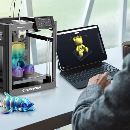 A person uses a tablet to control a FlashForge 3D Printer FlashForge Adventurer 5M - High-Speed Direct Extruder that is printing a colorful gradient teddy bear with high-performance 3D printing. A blue gradient dolphin figure is also present on the table, showcasing the printer's capabilities and high printing speed.