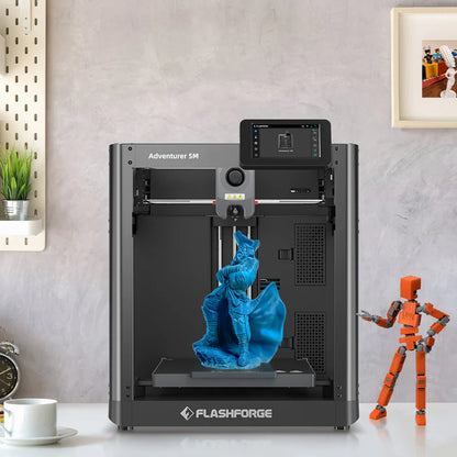 A 3D Printer FlashForge Adventurer 5M - High-Speed Direct Extruder on a desk, showcasing its high-performance 3D printing by producing a blue figurine. A small orange robot figurine stands to the right, while a mug and plant sit on the left side of the desk.