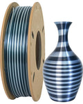 A spool of Toyar's PLA Silk Rainbow Colors Filament (1.75mm) beside a vase with metallic gray and black stripes, ideal for creating stunning 3D prints.