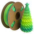 A spool of Toyar PLA Matte Rainbow 1.75mm Filament sits beside a green and yellow 3D printed Christmas tree topped with a star, highlighting the vibrant colors and smooth matte finish achievable with Toyar filament.
