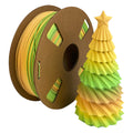A spool of Toyar PLA Matte Rainbow 1.75mm Filament with a matte finish sits beside a small, pleated yellow and green paper tree topped with a star.