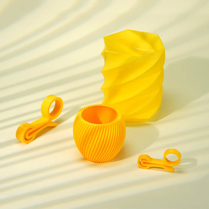 Four geometric yellow objects—printed with Flashforge PETG 1.75mm filament—include a wavy vase, ribbed sphere container, and two small clips, all arranged artfully on a light background with subtle shadows.