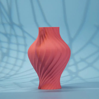 A coral-pink, matte vase with a spiral design—3D printed using Flashforge PLA 1.75mm Matte Colour filament from 3D Printing Perth - Cirrus Link—stands on a blue surface with crisscrossing shadow patterns in the background.