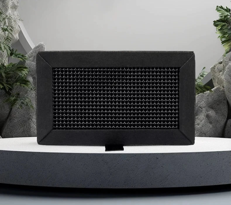 A black rectangular Bambu Lab X1/P1 Activated Carbon Air Filter with a textured front panel sits on a gray platform, subtly highlighted by green plants and rocks in the background. This sleek design, inspired by cutting-edge 3D printing techniques from Bambu Lab, seamlessly blends technology with nature while reducing odors and improving air quality.