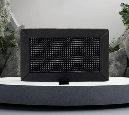 A black rectangular Bambu Lab X1/P1 Activated Carbon Air Filter with a textured front panel sits on a gray platform, subtly highlighted by green plants and rocks in the background. This sleek design, inspired by cutting-edge 3D printing techniques from Bambu Lab, seamlessly blends technology with nature while reducing odors and improving air quality.