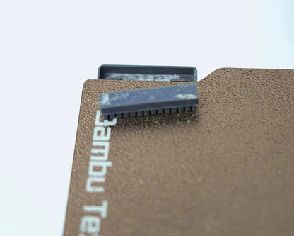 A close-up of a textured surface reveals the edge of a small, rectangular component from the Silicone Nozzle Wiper Brush Set (3-Pack) by Bambu Lab, crucial for maintaining the A1 Mini 3D Printer.