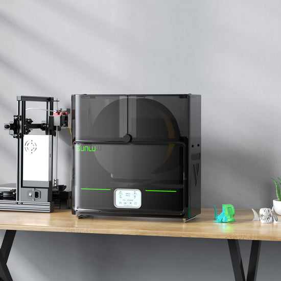 A 3D printer and the SUNLU S4 FilaDryer: 4-Bay Filament Drying Solution by SUNLU rest on a wooden table. A small green model stands beside the printer, which specializes in multi-color printing, while a potted plant decorates the scene.